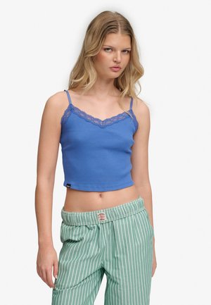 Młoda kobieta ubrana w niebieską koszulkę wykończoną koronką i zielone spodnie w paski, stojąca na jednolitym białym tle.