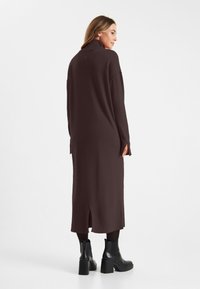 Lange, donkerbruine gebreide jurk met een hoge kraag, zijslits en ribgebreide accenten. Gekozen in combinatie met zwarte enkellaarzen.
