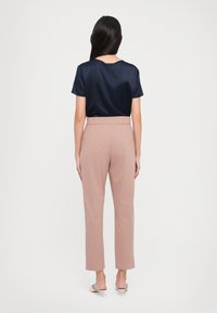 Donna in piedi di spalle, che indossa una blusa blu navy a maniche corte, pantaloni mauve a vita alta e mules bianchi con tacco basso su uno sfondo bianco.