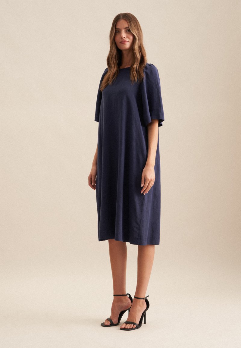 Navyblaues Leinenkleid mit lässigem Schnitt, kurzen Flatterärmeln und knielanger Länge. Getragen mit schwarzen Riemchen-High Heels.