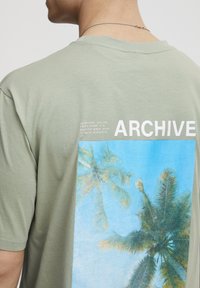 Oliņzaļš īsās piedurknes T-krekls, izgatavots no mīkstas auduma, ar zilu palmu koku grafiku uz muguras un baltu tekstu "ARCHIVE".