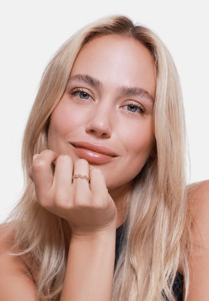 Ein roségoldener Zickzackring, der an einer Hand mit heller Haut und langen blonden Haaren getragen wird; der sanfte Hintergrund hebt das zarte Design des Rings hervor.