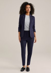 Donna con capelli ricci, in giacca e pantaloni blu navy, maglietta grigia chiara e mocassini neri, in piedi di fronte a uno sfondo beige.