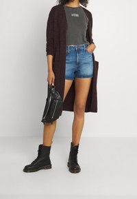 Mujer con un cárdigan burdeos oscuro, camiseta gris de corte corto con texto, pantalones cortos de mezclilla, botas negras con cordones, sosteniendo un bolso de mano de cuero negro.