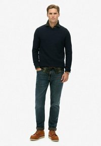 Marineblauer Strickpullover über einem grün-karierten Hemd, kombiniert mit blauen Jeans und braunen Schnürstiefeln. Das Model steht mit den Händen in den Taschen.