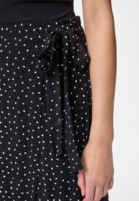 Melnās punktiņu svārki ir ar auduma jostu viduklī, tiem ir gluda tekstūra un visā virsmā ir baltas, vienmērīgi izvietotas punktiņas.