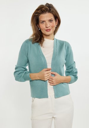 Frau mit braunen Haaren, die Perlenohrringe trägt, eine hellblaue gerippte Strickjacke über einem beigen Top und weiße Hose vor einfarbigem Hintergrund.