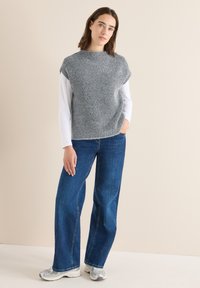 Grijs gestructureerd topje met korte mouwen gedragen over een wit shirt met lange mouwen, gecombineerd met wijde blauwe jeans en zilveren sneakers.