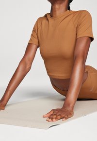 Person wearing brown activewear stretching on a beige yoga mat, supporting their upper body with extended arms.