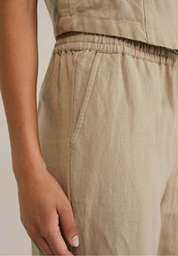 Pantaloni di lino beige con elastico in vita, dotati di tasche laterali e tessuto strutturato. Primo piano sulla vita e sulla mano.