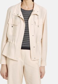 Jasnobeżowa kurtka o luźnym kroju, z dwoma przednimi kieszeniami, metalowymi zatrzaskami i ozdobnymi sznurkami. Nosi się ją na czarno-białą wzorzystą bluzkę.