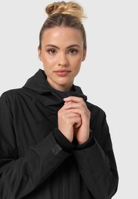 Black hooded jacket with a zip closure, ribbed cuffs, and a smooth texture. Model holds hands close together, showcasing the jacket's design.