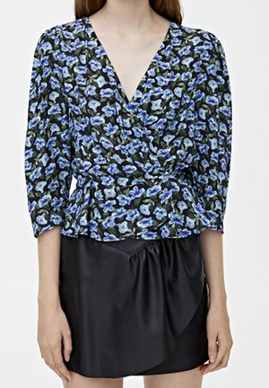 Femme portant un chemisier cache-cœur noir à imprimé floral bleu et une jupe asymétrique en cuir noire, montrée du cou à mi-cuisse.