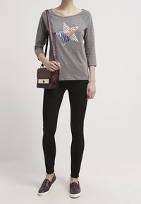 Graues Langarmshirt mit einem mehrfarbigen Sternmotiv, schwarze Leggings und einer dunkelmaronen Umhängetasche. Die Schuhe sind dunkelmarone Slipper.