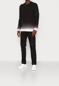 Pull en tricot noir avec un dégradé ombré vers le blanc, détails de poches sur les côtés, associé à un pantalon noir et des baskets blanches sur un fond neutre.