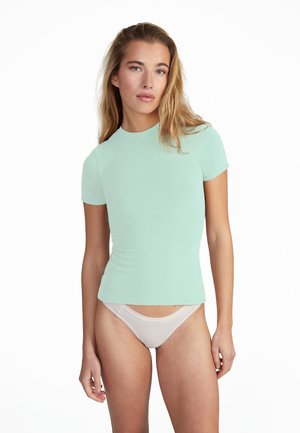 Junge Frau mit langen blonden Haaren, die ein hellgrünes, eng anliegendes Kurzarmshirt und weiße Unterwäsche vor einem weißen Hintergrund trägt.