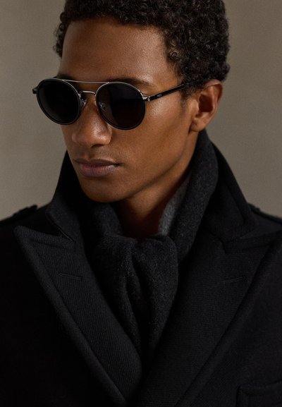 Occhiali da sole rotondi neri con lenti scure, montatura in metallo e design sottile; indossati con un cappotto nero e una sciarpa di lana scura.