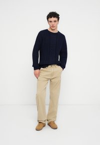 Πουλόβερ μπλε ναυτικού πλεξίματος, μπεζ παντελόνια άνετης εφαρμογής και καφέ σουέτ παπούτσια. Το μοντέλο στέκεται με τα χέρια στις τσέπες, σε ουδέτερο φόντο.