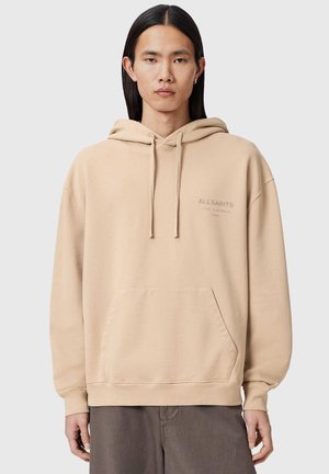 Junge Person mit langen schwarzen Haaren trägt einen beigefarbenen AllSaints-Kapuzenpullover mit Fronttasche und Kordeln und steht vor einem schlichten hellen Hintergrund.