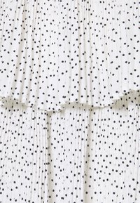 Tissu blanc à pois noirs irréguliers, avec une texture superposée plissée et des plis doux formant des lignes verticales.