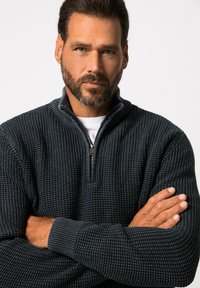 Dunkelblauer Strickpullover mit einem halben Reißverschluss, strukturiertem Muster und gerippten Bündchen. Das Model posiert mit verschränkten Armen vor einem schlichten Hintergrund.