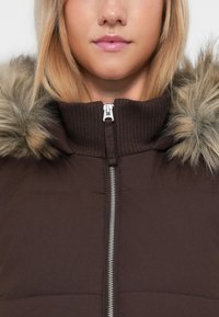 Detailní záběr na osobu v hnědé zipové bundě s vroubkovaným límcem a kapucí s kožešinovým lemem, viditelné rty s jemným růžovým leskem.