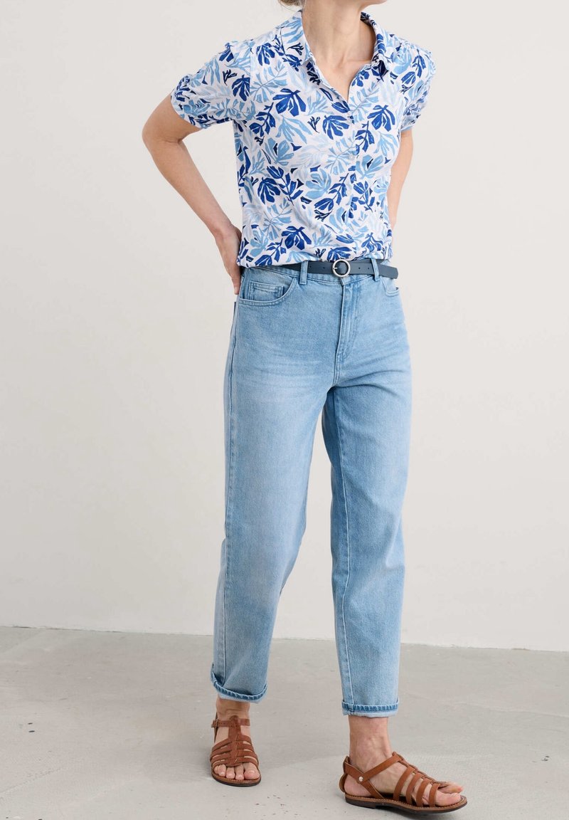 Femme portant un chemisier à manches courtes fleuri bleu clair, un jean bleu clair taille haute avec une ceinture noire, et des sandales marron à brides, debout les mains sur les hanches.