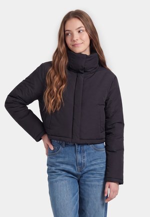 Junge Frau mit langen Haaren, die eine schwarze Steppjacke und blaue Jeans trägt, steht mit einer Hand in der Hüfte und schaut zur Seite.