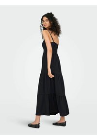 Robe maxi noire avec des bretelles fines et un corsage smocké. Présente des couches en tiers et une silhouette fluide. Portée avec des chaussures plates à clous.