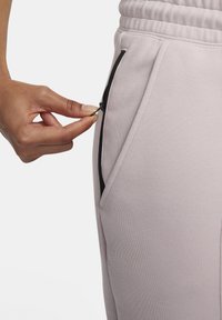 Pantaloni della tuta rosa chiaro con una texture liscia, dotati di una tasca con zip nera. Vita elastica visibile con dettagli di cucitura.