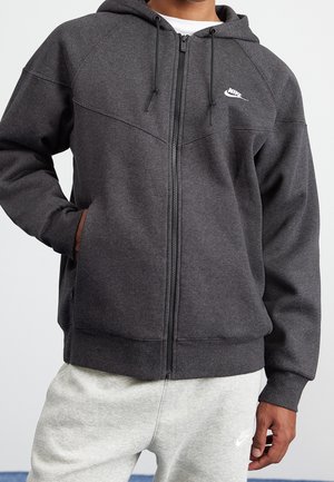 Person iført mørkegrå Nike hættetrøje med lynlås og snore samt lysegrå Nike joggingbukser, stående med den ene hånd i lommen.