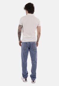 Hombre con cabello rizado y tatuajes, vestido con una camiseta polo blanca, jeans azules y zapatillas blancas, de pie y de espaldas.