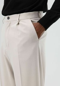 Crèmekleurige broek met een gladde textuur, voorzien van een voorplooi, knoopsluiting en zijzakken. Gecombineerd met een zwart topje met lange mouwen.