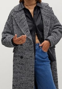 Person in einem schwarz-weißen Fischgrät-Mantel, schwarzem Lederhemd, beigem Rollkragenpullover, blauen Jeans und goldenen Ringen an beiden Händen.