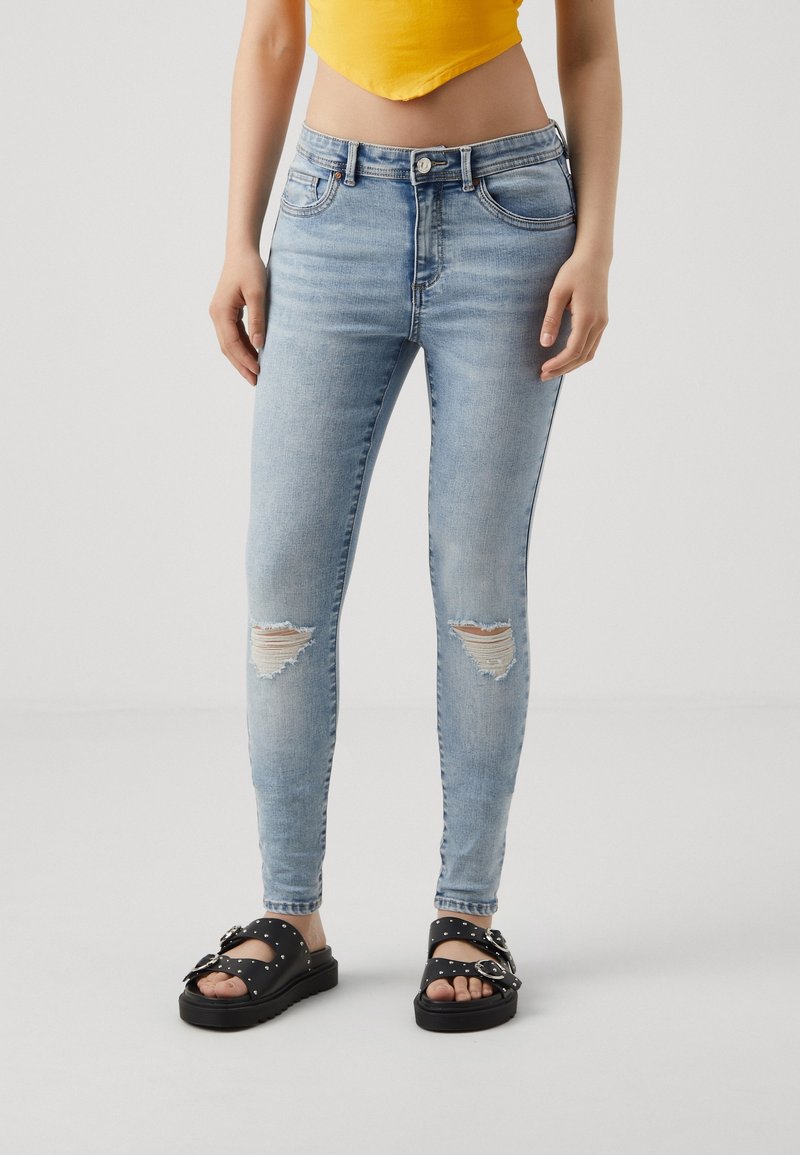 Personne portant un jean skinny déchiré bleu clair, un top court jaune et des sandales noires cloutées, debout sur un fond uni.