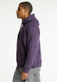 Sudadera morada con cordón ajustable, puños y dobladillo acanalados, corte suelto, material de algodón; combinada con jeans gris claro.