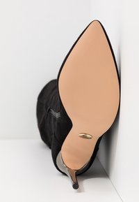 Chaussure à talon haut en daim noir avec un bout pointu et une semelle beige lisse. Présente des accents métalliques près du talon et un détail de fermeture éclair sur le côté.