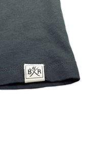 Dunkelgrauer Baumwollstoff mit glatter Textur; am Saum befindet sich ein gewebtes Etikett mit schwarzem Text und gekreuztem Design.