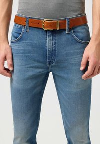 Jeans en denim bleu avec une finition délavée, portés avec une ceinture en cuir beige dotée d'une boucle dorée et d'une étiquette de marque sur le côté.