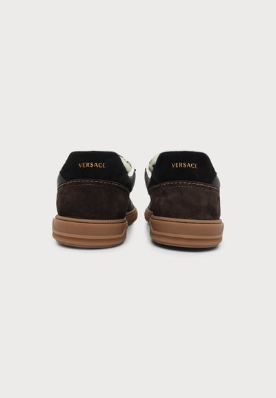 Paire de baskets marron et noir avec des semelles en caoutchouc couleur beige, vues de dos, chaque talon avec le logo doré "Versace".
