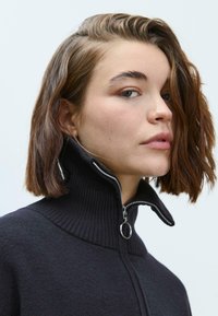 Pull noir zippé avec col côtelé et quincaillerie en argent, présentant une texture douce et un design ajusté.
