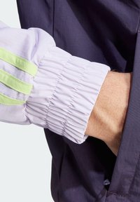 Chaqueta morada con puños de lilas fruncidos que presentan tres rayas verdes. La tela parece suave con detalles de costura visibles.