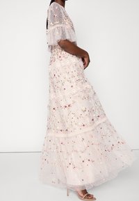 Femme portant une robe longue transparente avec broderies florales et détails en dentelle, associée à des sandales à talons hauts couleur nude.
