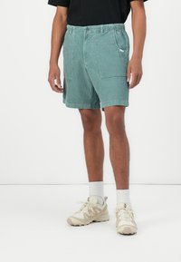 Green denim shorts with an elastic waistband, two front pockets, and a side pocket. Paired with a black shirt and light shoes.