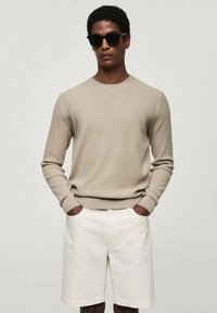 Beige ribbed knit sweater with a crew neckline, paired with light-colored shorts. Model wears black sunglasses. Smooth texture.