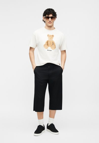 Homme portant des lunettes de soleil, un t-shirt blanc avec un motif d'ours en peluche, un pantalon noir court, des chaussettes blanches et des baskets noires, debout les mains dans les poches.