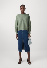 Svetlo zelena pulover z napihnjenimi rokavi, kombiniran z temno jeans krilom, ki ima srednji razporek. Drži majhno svetlo modro torbico. Črne sandale na blok peto.