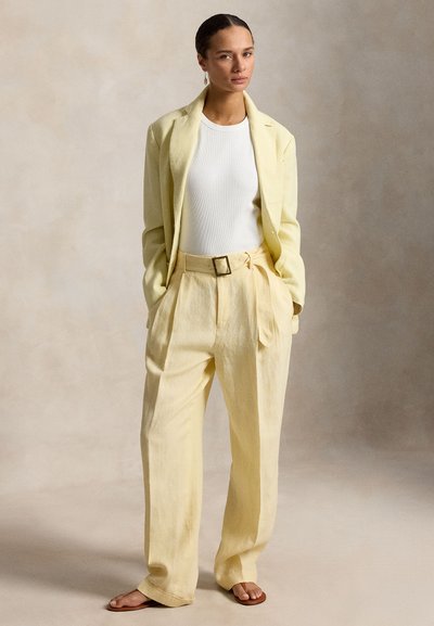 Γυναίκα με τα μαλλιά μαζεμένα προς τα πίσω, φορώντας ένα ανοιχτό κίτρινο κοστούμι με φαρδιές παντελόνες και λευκό τοπάκι, στέκεται με τα χέρια στις τσέπες μπροστά σε ουδέτερο φόντο.