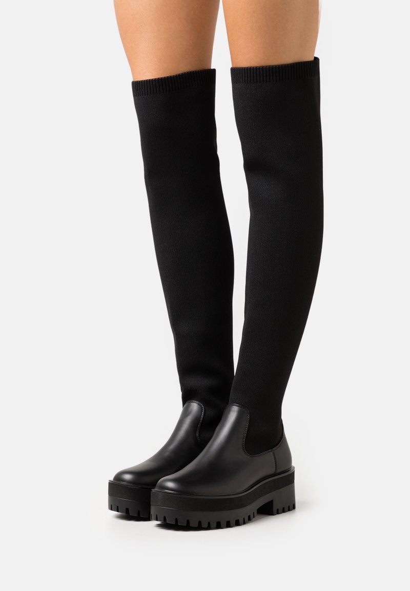 Bottes noires montantes jusqu'aux genoux avec semelles épaisses à crampons, portées par une personne debout devant un fond clair uni.