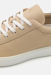 Sneaker in pelle beige con lacci bianchi, texture liscia e suola in gomma bianca a contrasto. Presenta un design minimalista e cuciture sottili.